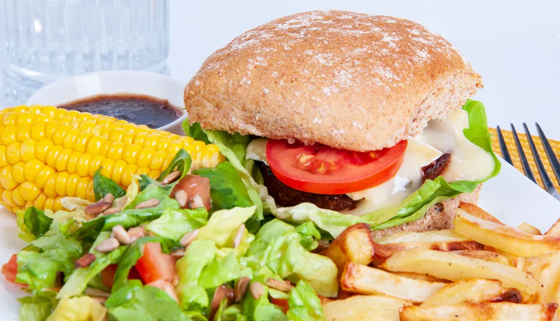 Op een bord een vegetarische hamburger met gesmolten brie, een maiskolf, salade, zonnebloempitten en zelfgemaakte frietjes