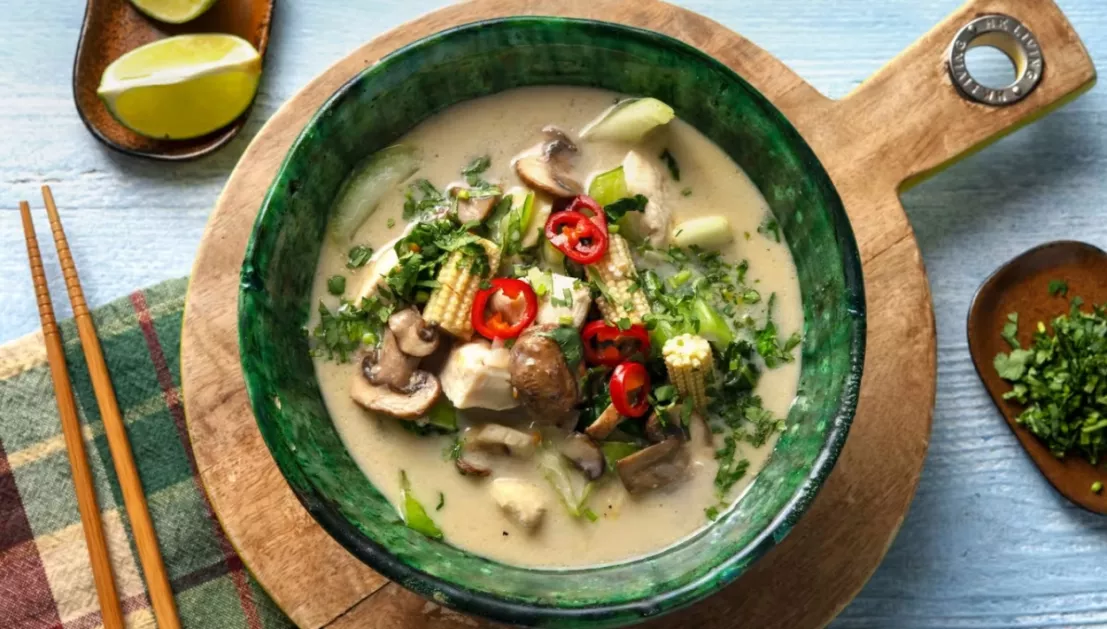 Tom kha kai soep met champignons en mais geserveerd in een soepkom met daaronder een ronde houten plank