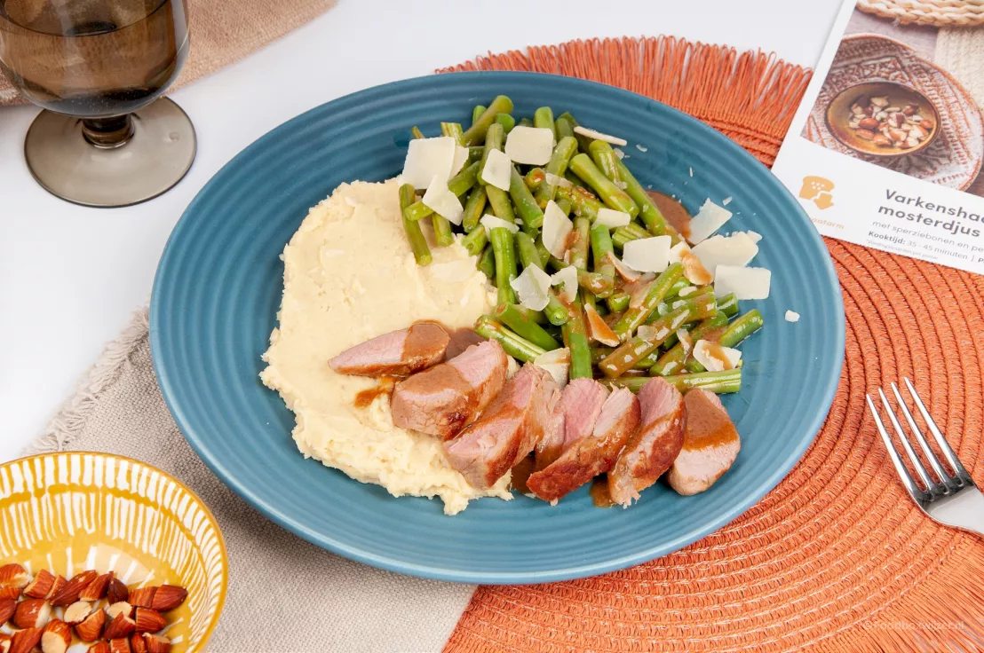 Op een blauw bord met placemat eronder een gerecht van green chef met varkenshaas, puree, sperziebonen en zelfgemaakte honing mosterd jus